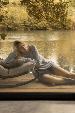 Woman reclining in tranquility wearing MANITO's Platinum Blue Wunder Silk Wool Cashmere Tracksuit, with a reflective water backdrop.