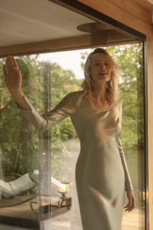 Woman in a coral gold Muses Silk Midi Dress by MANITO, greeting the morning light with a serene smile, in a harmonious indoor-outdoor space.