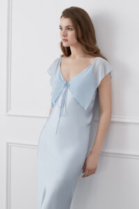 Woman in a Crete Silk Midi Dress in blue with fluttering ruffle details and drawstring neckline for a chic, airy look.