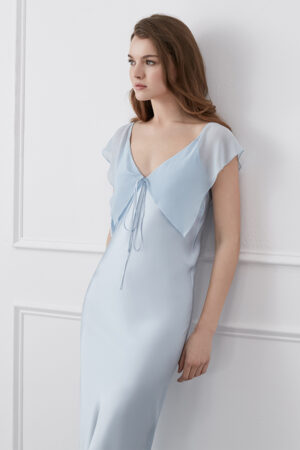 Woman in a Crete Silk Midi Dress in blue with fluttering ruffle details and drawstring neckline for a chic, airy look.