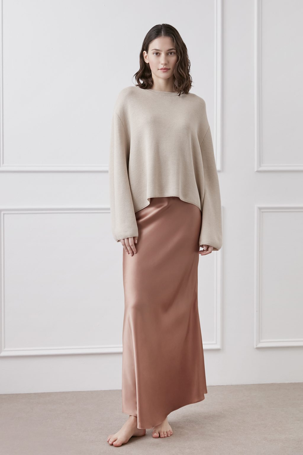 Confident woman in an ivory cashmere top with MANITO's coral gold Muses Silk Midi Skirt, poised in a minimalist setting, exuding sophistication.