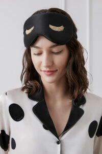 Woman wears the Lash Contour Silk Eye Mask in black, showing the elegant gold embroidery and smooth silk finish.