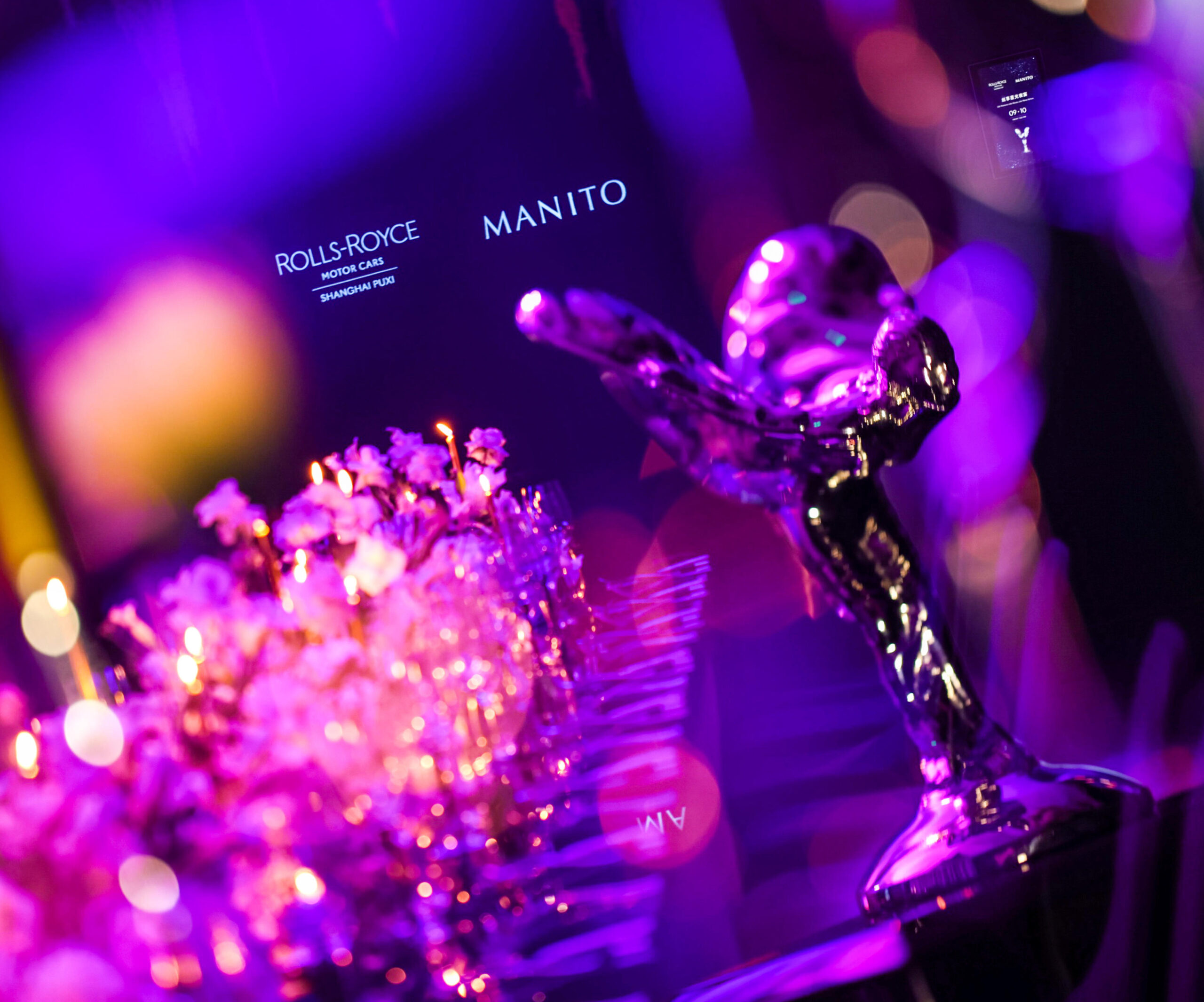 An exclusive event showcasing the partnership between MANITO and Rolls-Royce, highlighting the iconic Rolls-Royce hood ornament with MANITO's branding in the backdrop, symbolizing a fusion of luxury automotive excellence and refined silk elegance.