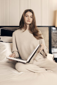 Thoughtful woman enjoying a book, dressed in MANITO's sand-colored Silk Wool Cashmere Loungewear Set, in an elegant bedroom setting.