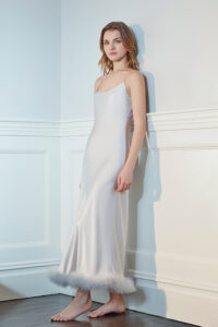 Elegant woman in a Diva Feather Trimmed Long Silk Slip Dress standing against an ornate wall, showcasing grace and luxury.