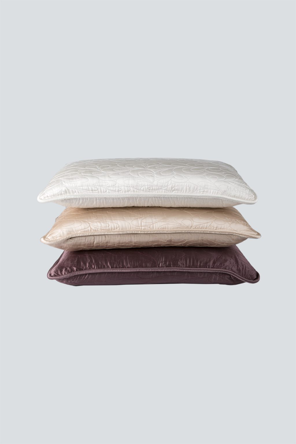 A stack of quilted lumbar silk pillows in varying shades, showcasing the MANITO brand embroidery.
