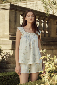 Woman wearing a silky floral short pajama set, standing in a lush garden with classical architecture in the background.