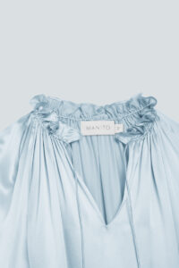 Close-up of ruffle trim neckline and MANITO logo on blouse, showcasing high-quality craftsmanship and strong gloss.