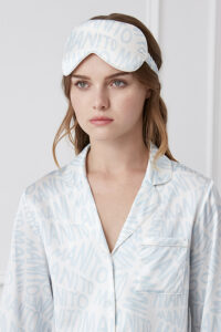 Woman wearing a white silk eye mask with blue 'MANITO' script pattern matching her pajamas, looking serene.