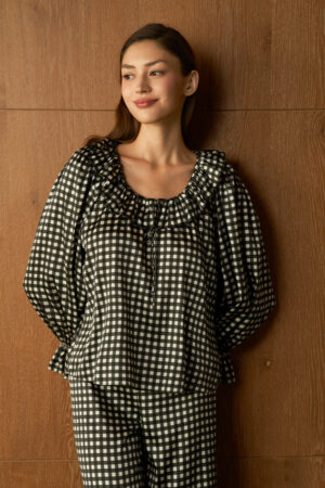 Woman in black Blooming Women's Pajama Set, leaning against a wooden wall, smiles gently in warm light.