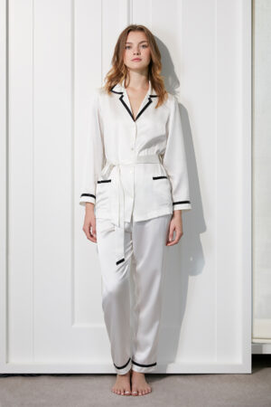 Woman in Coco Lady Silk Pajama Set with classic white with black piping detail, featuring a button-up top with a notched collar.