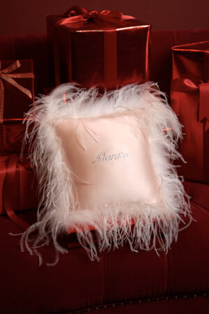 Pink holiday throw pillow adorned with delicate white feathers and rhinestones, paired with festive gift boxes.