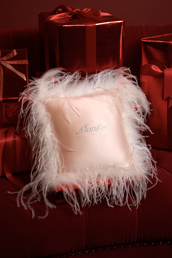Pink holiday throw pillow adorned with delicate white feathers and rhinestones, paired with festive gift boxes.