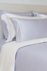 Side view of MANITO Fluora Silk Pillowcase Set on a bed, showing the contrast color borders and lustrous silk finish in lavender and white.