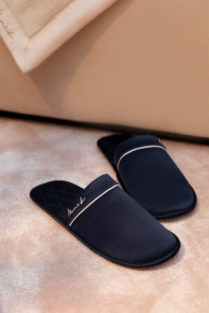 A pair of luxurious men's silk lounge shoes with elegant white detailing, placed beside a soft beige cushion.