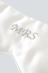 A close-up of an elegant white silk eye mask featuring a "MRS" monogram in beautiful gold embroidery, showcasing exquisite detail and sophistication.