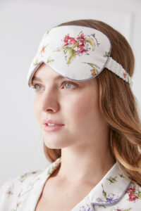Woman wears a Lrisette silk eye mask adorned with a vivid floral design, showcasing soft pastel colors and intricate flower illustrations, adding a delicate and elegant touch.