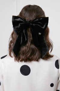 Stylish woman wearing a Duchess Silk-Velvet Bow Barrette on her hair, detailed with a monogrammed 'M', set against a polka dot sweater.