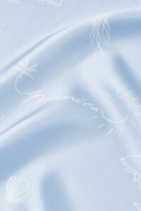 Close-up of MANITO Ocean Blue Silk Scarf highlighting brand signature and sun sketch on silky fabric.