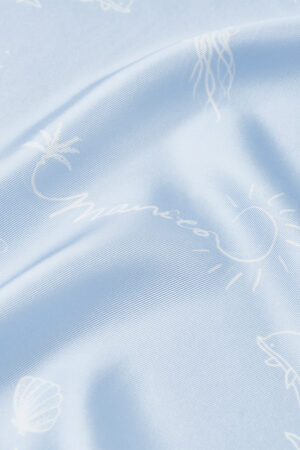 Close-up of MANITO Ocean Blue Silk Scarf highlighting brand signature and sun sketch on silky fabric.