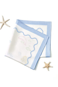 Folded MANITO Ocean Wave Silk Scarf with pastel sea motifs and soft blue border, styled with starfish decor.