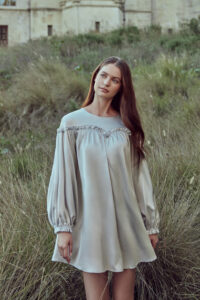 Woman in silver gray silk mini dress with lantern sleeves and unique ruffle collar detail, standing against natural background with grass.