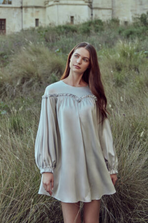 Woman in silver gray silk mini dress with lantern sleeves and unique ruffle collar detail, standing against natural background with grass.