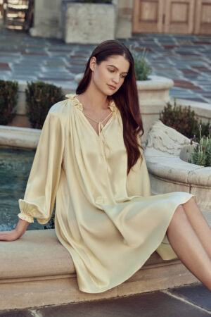 A woman wearing an elegant bellflower yellow silk shirt dress adorned with ruffle trim and lantern sleeves.
