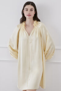 Smiling woman wearing bellflower yellow silk dress features ruffle trim collar, leaning against white wall.