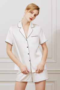 Woman in elegant Treasure short pajama set in ivory silk with black contrast piping.