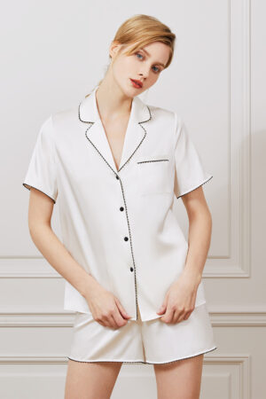 Woman in elegant Treasure short pajama set in ivory silk with black contrast piping.