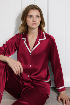 Seated model in burgundy Coco Lady Pajama Set, showing glossy finish and sharp contrast piping.