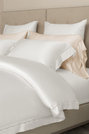 Elegant white and champagne-colored MANITO New Aqua silk bedding set featuring delicate detailing, displayed on a modern bed.