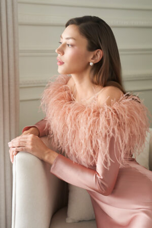 Woman leans on an armchair in coral New Diva Feather Off-the-shoulder Dress, feather-trimmed neckline glowing softly in light.