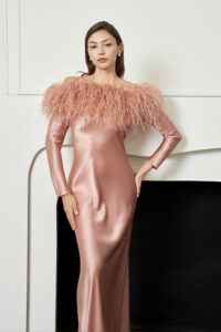 Woman stands by a fireplace in coral New Diva Feather Off-the-shoulder Dress, showing long satin skirt and feather neckline.