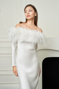 Woman poses in white New Diva Feather Off-the-shoulder Dress, with delicate feather trim framing the neckline for a romantic look.