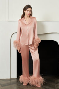 Woman stands by the fireplace in coral New Diva Feather Pajamas Set, satin top and feather-trimmed trousers creating modern loungewear.