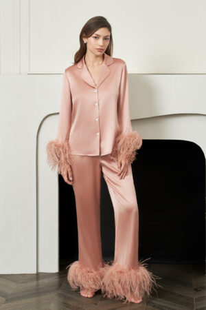 Woman stands by the fireplace in coral New Diva Feather Pajamas Set, satin top and feather-trimmed trousers creating modern loungewear.