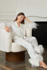 Smiling woman relaxes in white New Diva Feather Pajamas Set, feather-trimmed details bringing dreamy charm to everyday lounging.