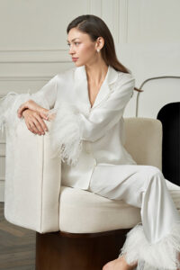 Woman sits sideways in white New Diva Feather Pajamas Set, soft feathers at the cuffs resting on the chair for quiet evening comfort.