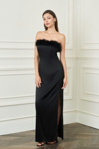 Woman stands in black New Diva feather strapless dress, sleek column silhouette with thigh slit, adding subtle drama in a classic room.