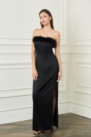 Woman stands in black New Diva feather strapless dress, sleek column silhouette with thigh slit, adding subtle drama in a classic room.