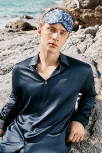 Model wearing MANITO Ocean Blue Dolphin Silk Eye Mask by the rocky seaside in a navy silk shirt.