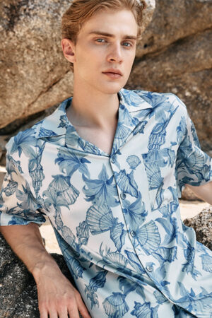 A man reclines against a rock, wearing MANITO Ocean Blue Men's Silk Shorrt Pajama Set, adorned with an elegant blue floral print.