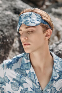 A man is closing his eyes by the sea, wearing MANITO Ocean Blue Men's Silk Eye Mask.
