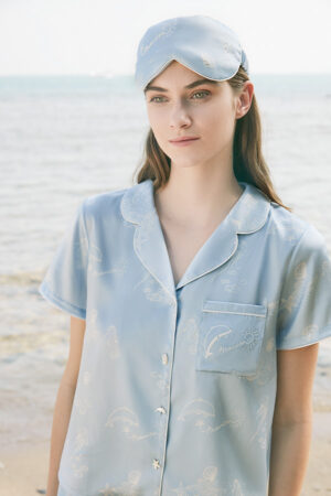 Woman wearing marine print pajama and eye mask from MANITO Ocean Blue Sleeping Beauty Gift Box, standing by the sea.