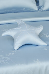MANITO blue Ocean Starfish Throw Pillow on silk bedspread with embroidered shells and sea creatures.