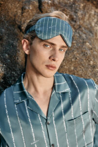 Model leaning against rock wearing MANITO Ocean Stripe Men's Silk Eye Mask and matching shirt.