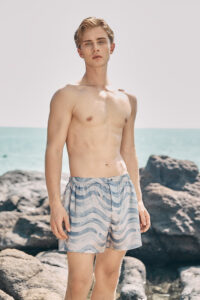 Model wearing MANITO Ocean Wave Men's Shorts, showcasing the full front design near the ocean.