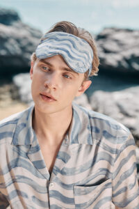 Model wearing the MANITO Ocean Wave Men's Silk Eye Mask and matching shirt while sitting on beach rocks.